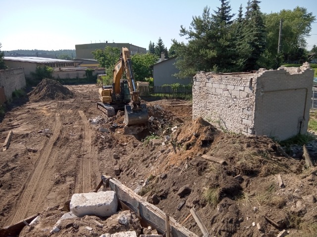 Żółta koparka na placu budowy, w tle resztki muru z cegły, widoczne ślady opon na ziemi.