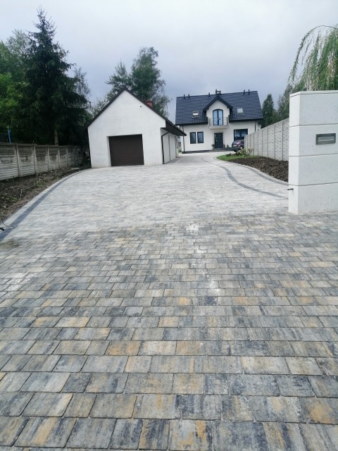 Szeroki, zakrzywiony podjazd z kostki brukowej w odcieniach szarości i beżu prowadzący do białego domu z czarnym dachem i garażu; widoczne świeżo posadzone rośliny wzdłuż obrzeży.