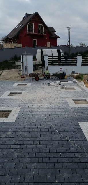 Częściowo ułożona nawierzchnia z kostki brukowej w odcieniach szarości z kwadratowymi wstawkami, widoczni dwaj pracownicy z taczkami i narzędziami w trakcie pracy, w tle czerwony dom i biały bus.