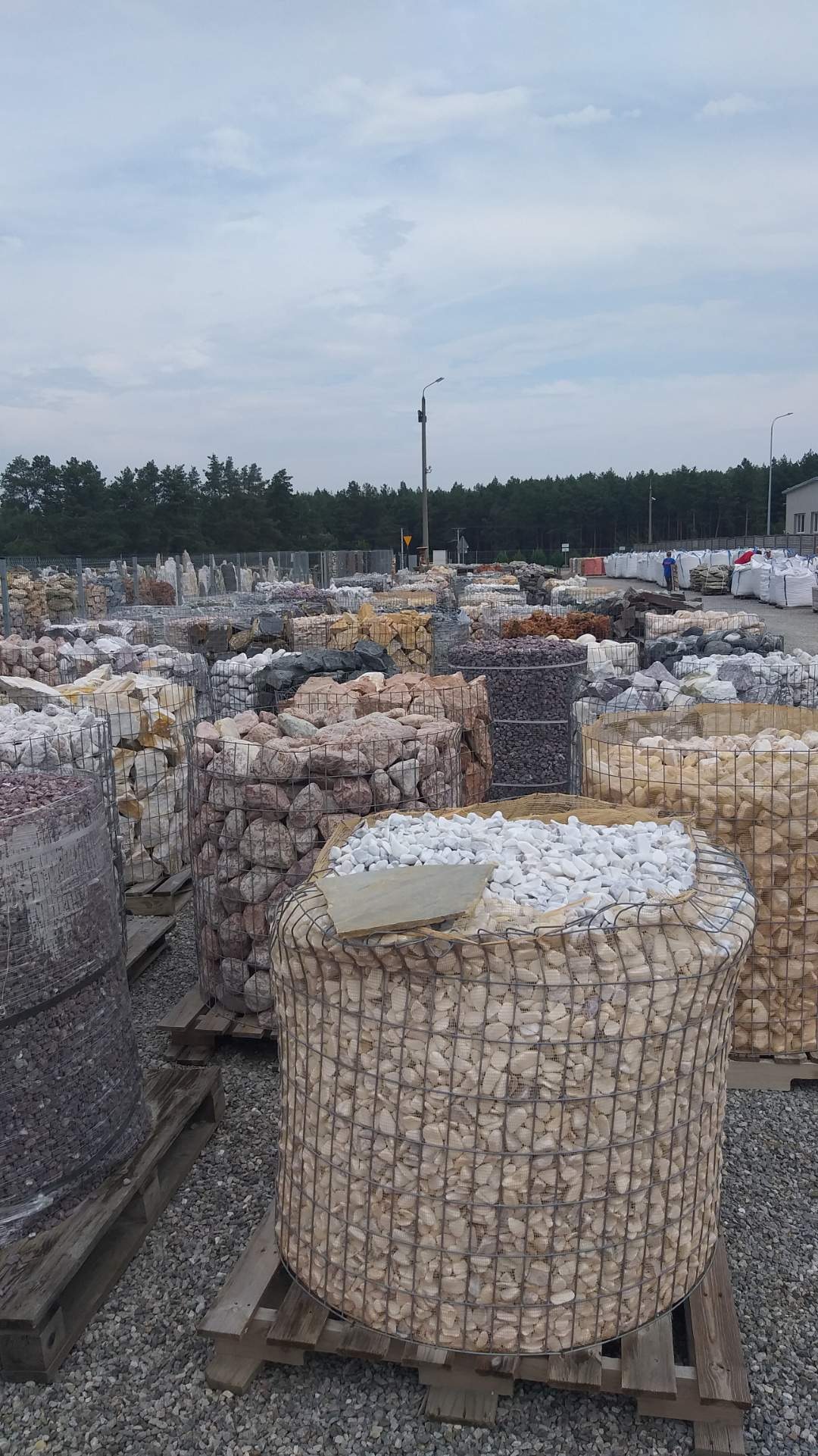 Szeroki plan składu z koszami gabionowymi wypełnionymi różnokolorowymi kamieniami dekoracyjnymi, niektóre z drewnianymi pokrywami, na tle lasu i budynku.