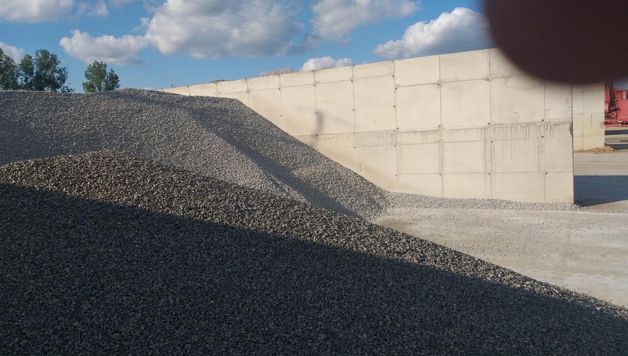 Góra szarego kruszywa na placu budowy, z betonową ścianą w tle i fragmentem nieba z chmurami