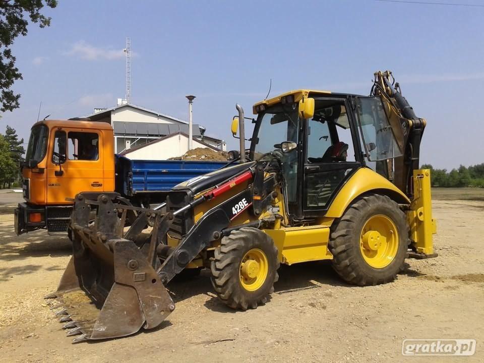 Żółto-czarna koparko-ładowarka 428E stoi obok pomarańczowej ciężarówki z niebieską skrzynią ładunkową na placu budowy; w tle budynek z anteną.