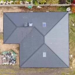 Widok z góry na ciemnoszary dach domu jednorodzinnego z dwoma oknami dachowymi i kominem, obok składowane materiały budowlane i resztki po budowie.