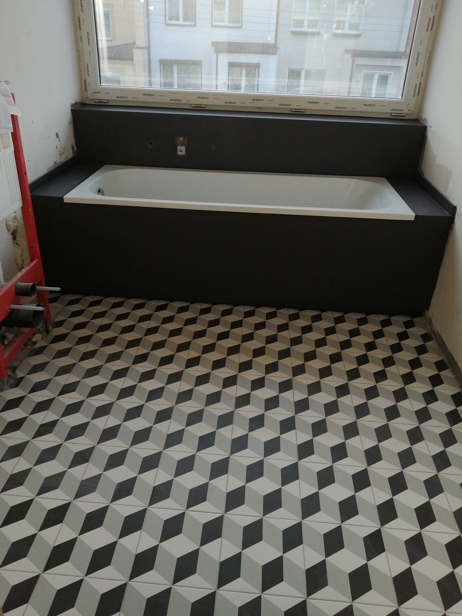 Biała wanna obudowana ciemnoszarą płytą w trakcie montażu, widoczne geometryczne płytki podłogowe w odcieniach szarości, fragment ściany z widocznym stelażem podtynkowym i rurami.