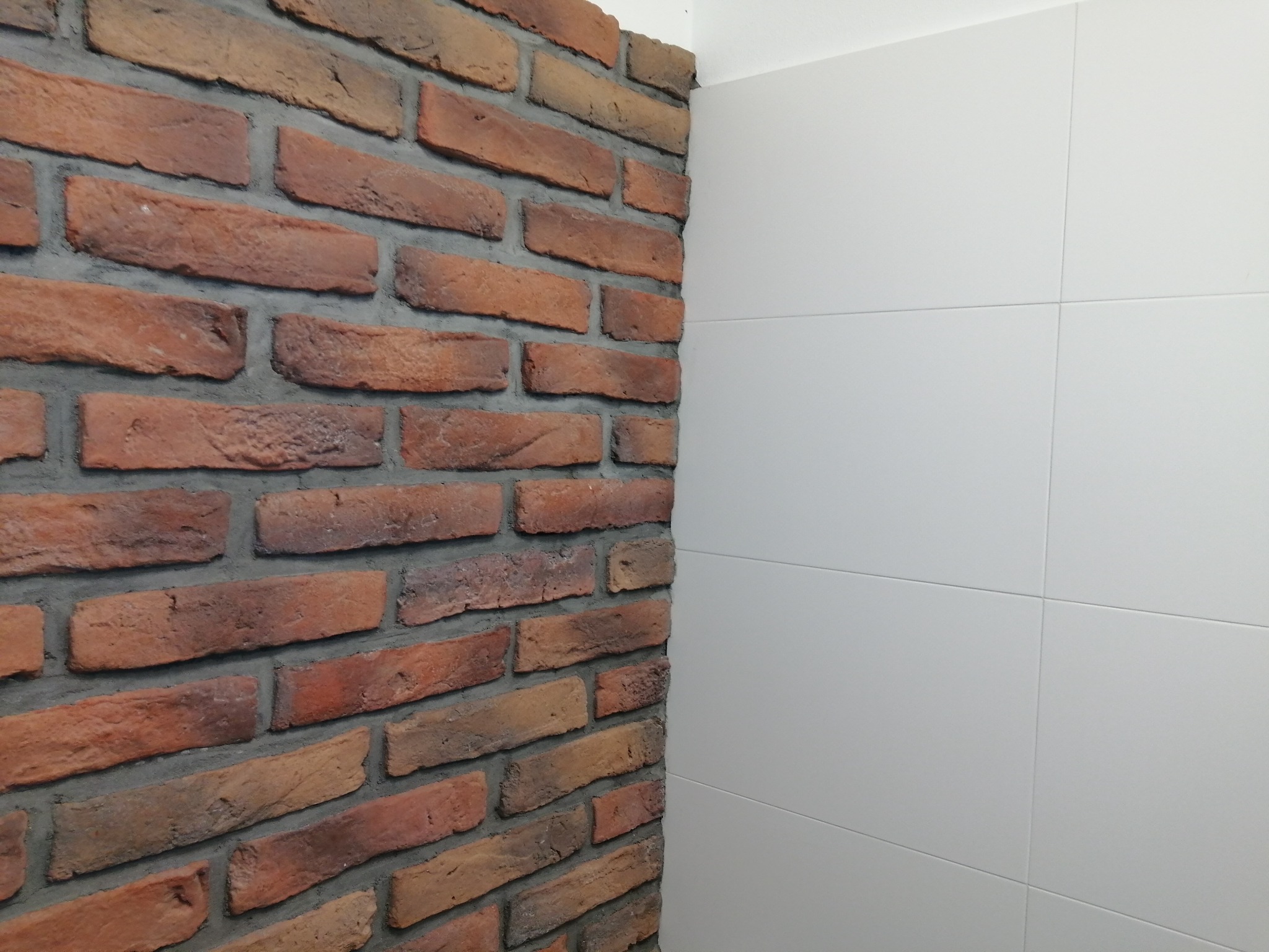 Fragment ściany wykończonej czerwoną cegłą klinkierową z szarą fugą, sąsiadujący z białą ścianą wyłożoną dużymi, matowymi płytkami ceramicznymi.