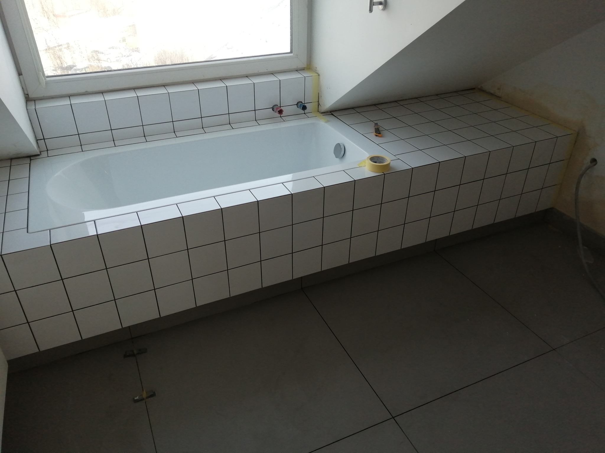 Biała wanna obudowana kwadratowymi płytkami w trakcie prac wykończeniowych w łazience na poddaszu, widoczne taśmy malarskie zabezpieczające krawędzie i krzyżyki dystansowe na podłodze.