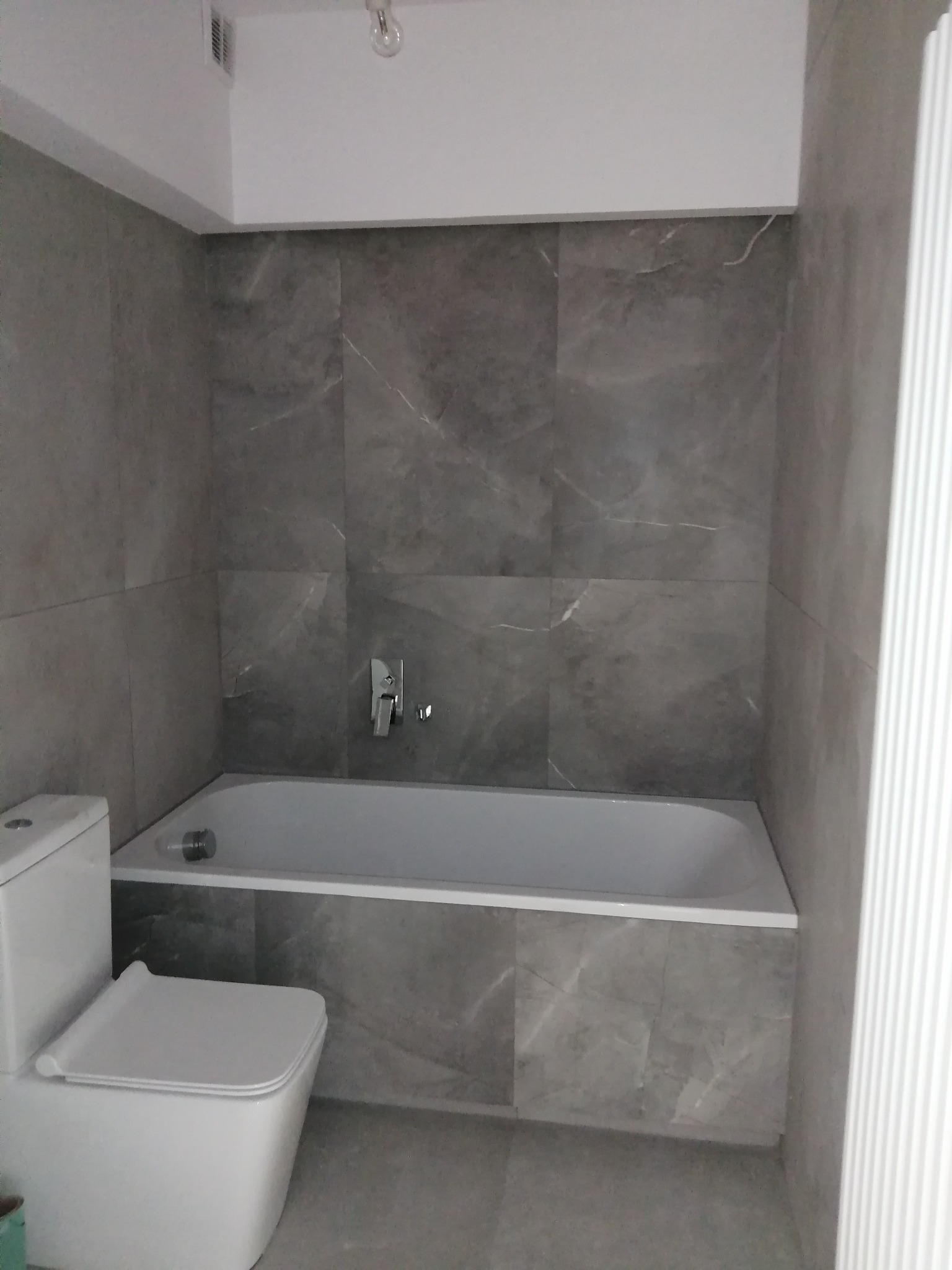 Wykończona łazienka z szarymi płytkami imitującymi marmur, biała wanna z baterią, toaleta kompaktowa z prostokątną miską oraz grzejnik żeberkowy.