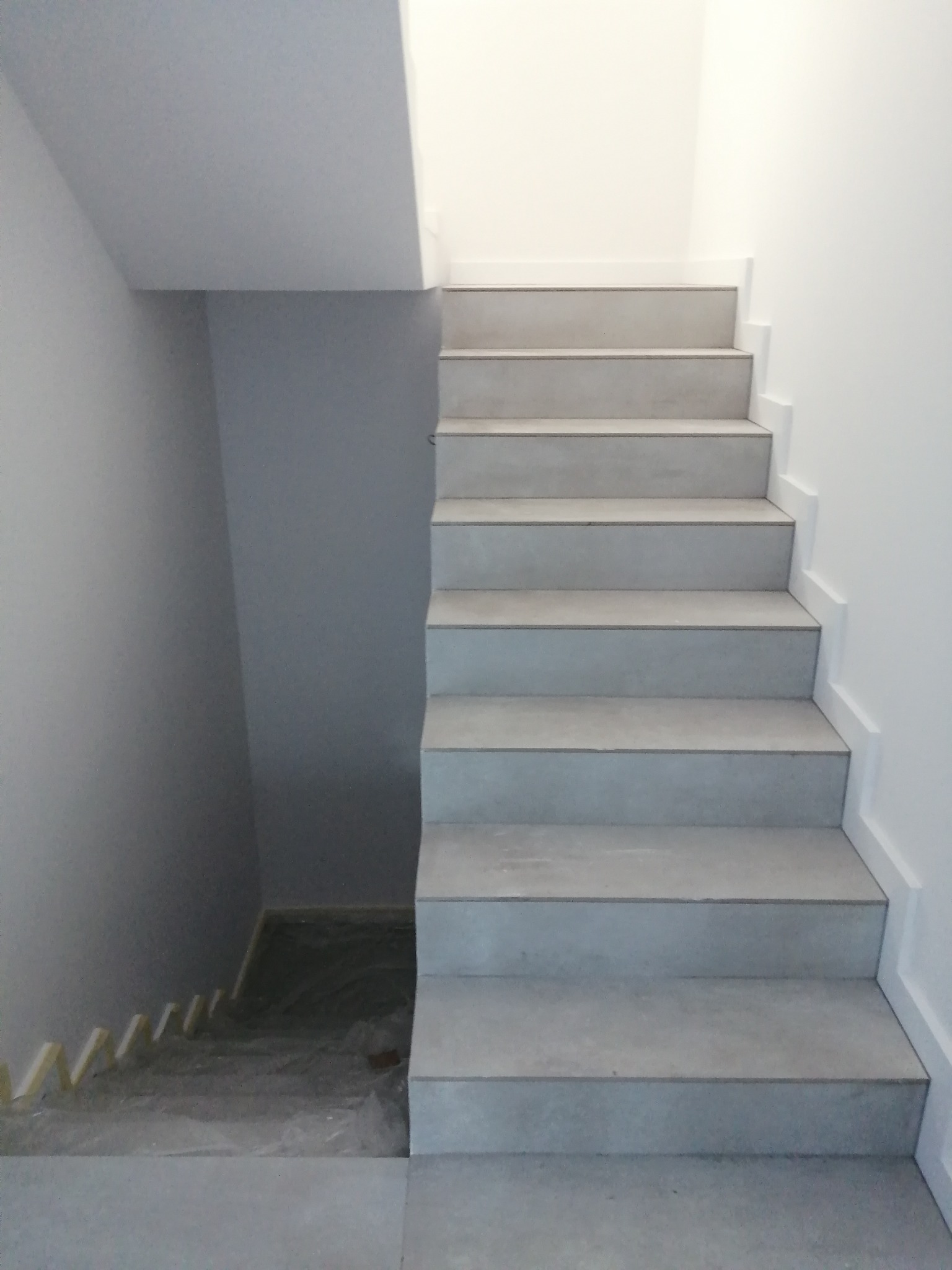Wykończone schody z szarych płytek, z widocznym zabezpieczeniem folią i listwami na dolnym biegu schodów, białe ściany.