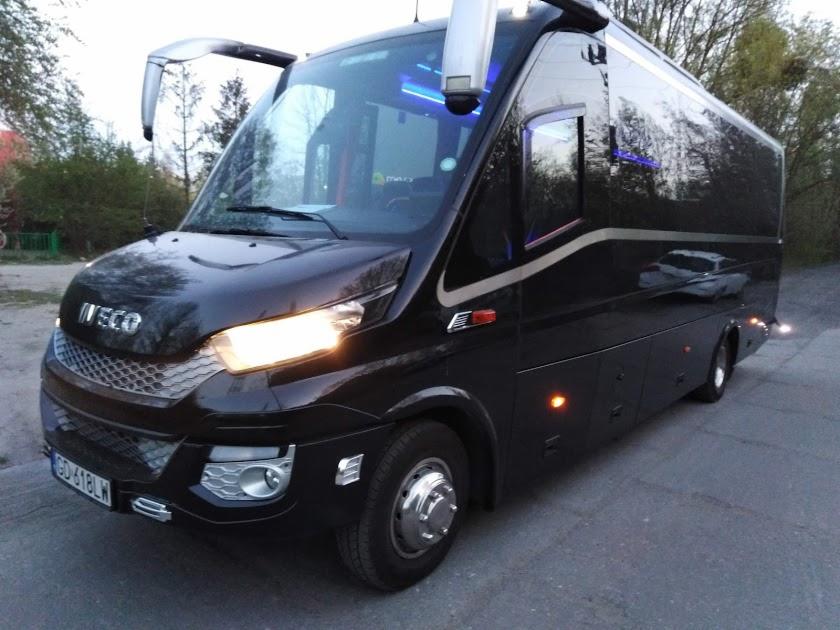 Czarny, luksusowy bus Iveco z włączonymi światłami mijania, zaparkowany na zewnątrz, z widocznym odbiciem samochodu w bocznej szybie.