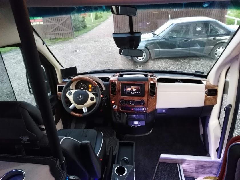 Wnętrze luksusowo wyposażonego busa z drewnianymi elementami wykończeniowymi deski rozdzielczej i jasną kierownicą, na zewnątrz zaparkowany ciemny samochód osobowy.