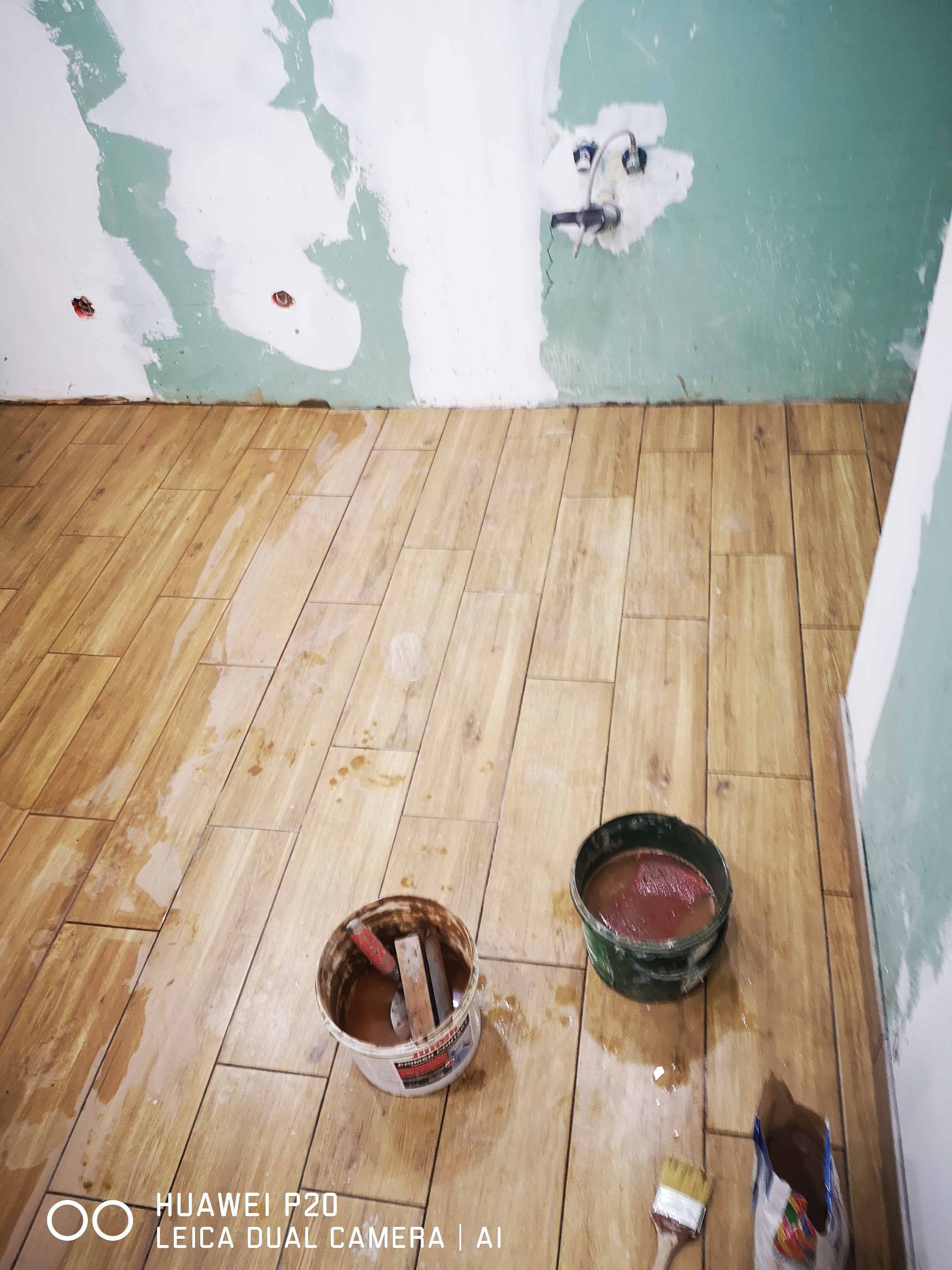 Układanie drewnopodobnych płytek na podłodze w trakcie remontu, widoczne dwa wiadra z zaprawą, pędzel i worek kleju.