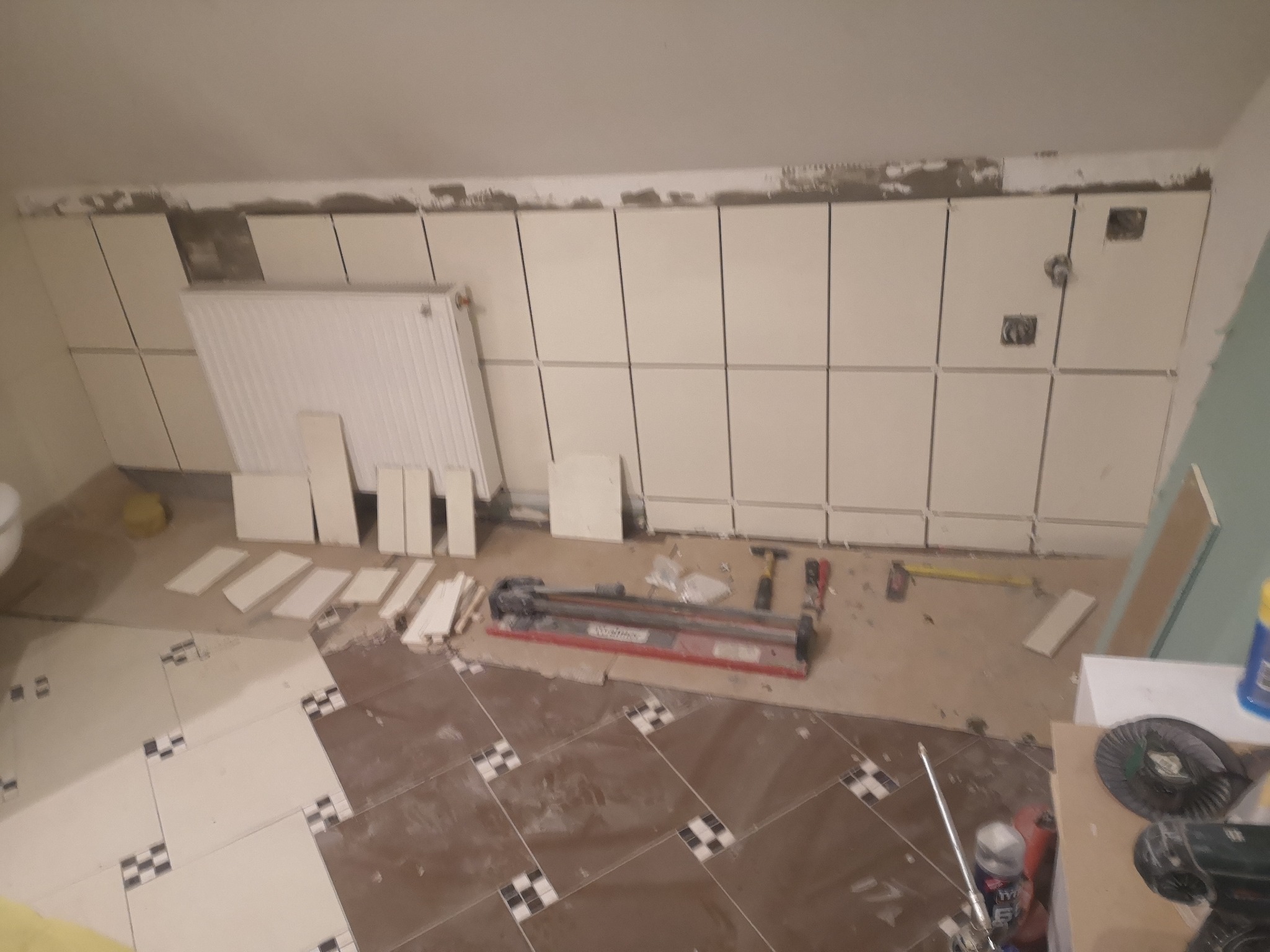Układanie białych płytek na ścianie w łazience na poddaszu, widoczne narzędzia, grzejnik i częściowo ułożona podłoga z brązowo-białych płytek.