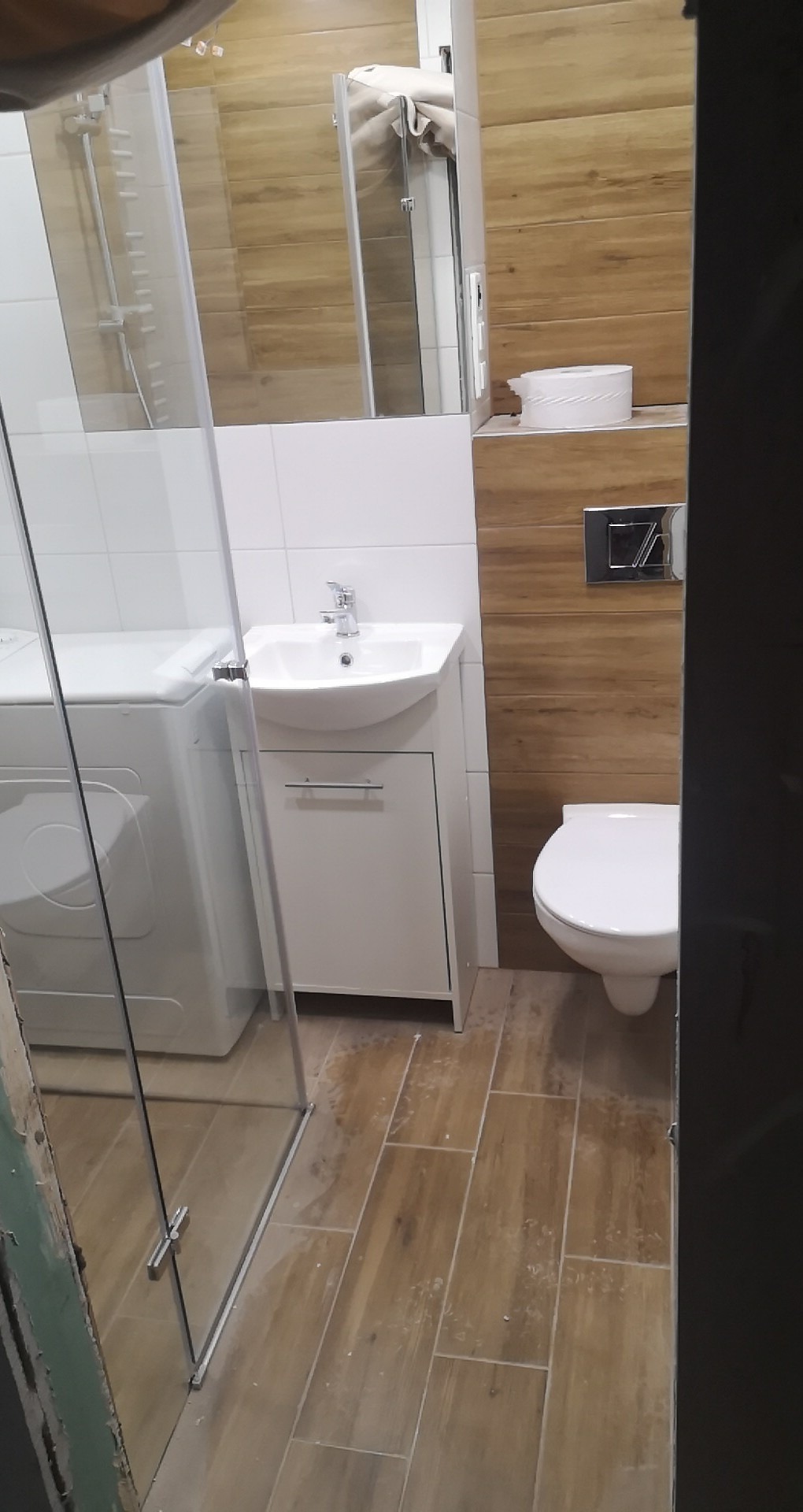 Ukończona łazienka z kabiną prysznicową, pralką, umywalką z szafką i toaletą, wykończona płytkami imitującymi drewno na ścianach i podłodze, widoczne ślady po remoncie na podłodze.