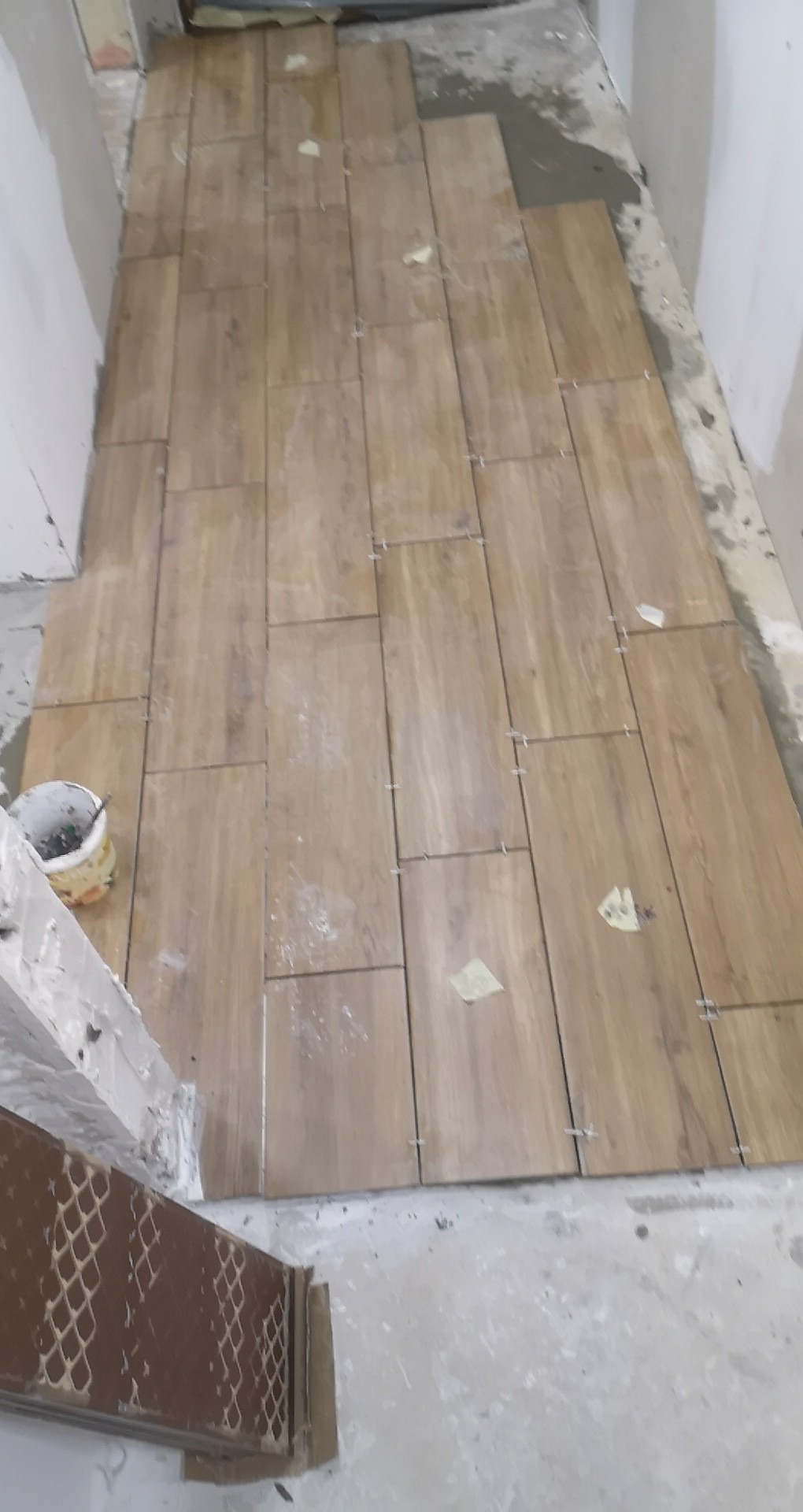 Układanie podłogi z płytek imitujących drewno, widoczne krzyżyki dystansowe i fragment wiaderka z klejem.