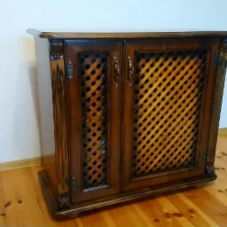 Ręcznie wykonana, ciemnobrązowa szafka z drewnianymi drzwiczkami z kratką, z ozdobnymi zawiasami i uchwytami, stojąca na drewnianej podłodze przy białej ścianie.