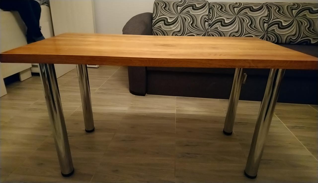 Prostokątny stół z blatem z jasnego drewna i chromowanymi nogami, ustawiony na podłodze z płytek imitujących drewno, w tle sofa z wzorzystą tapicerką.