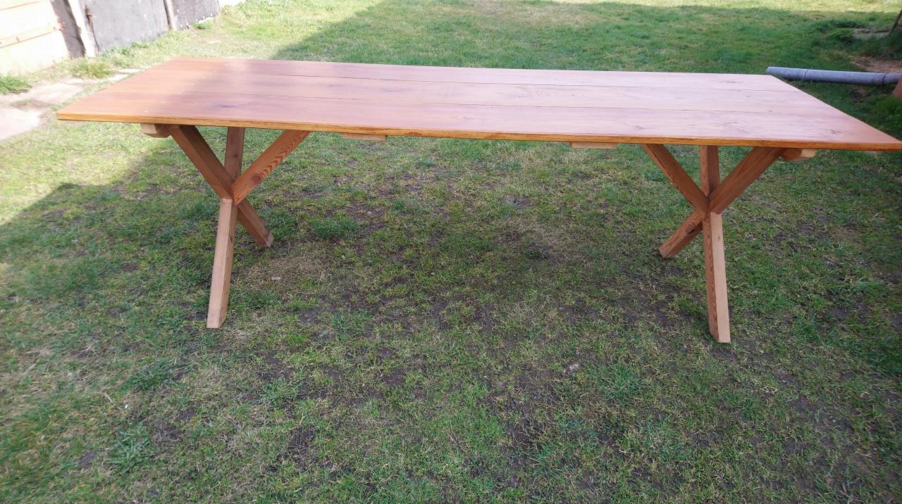 Rustykalny, drewniany stół ogrodowy z blatem z desek i nogami w kształcie litery X, ustawiony na trawniku.