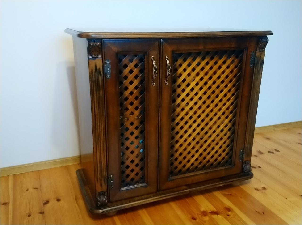 Ręcznie wykonana, ciemnobrązowa szafka z drewnianymi drzwiczkami z kratką, z ozdobnymi zawiasami i uchwytami, stojąca na drewnianej podłodze przy białej ścianie.