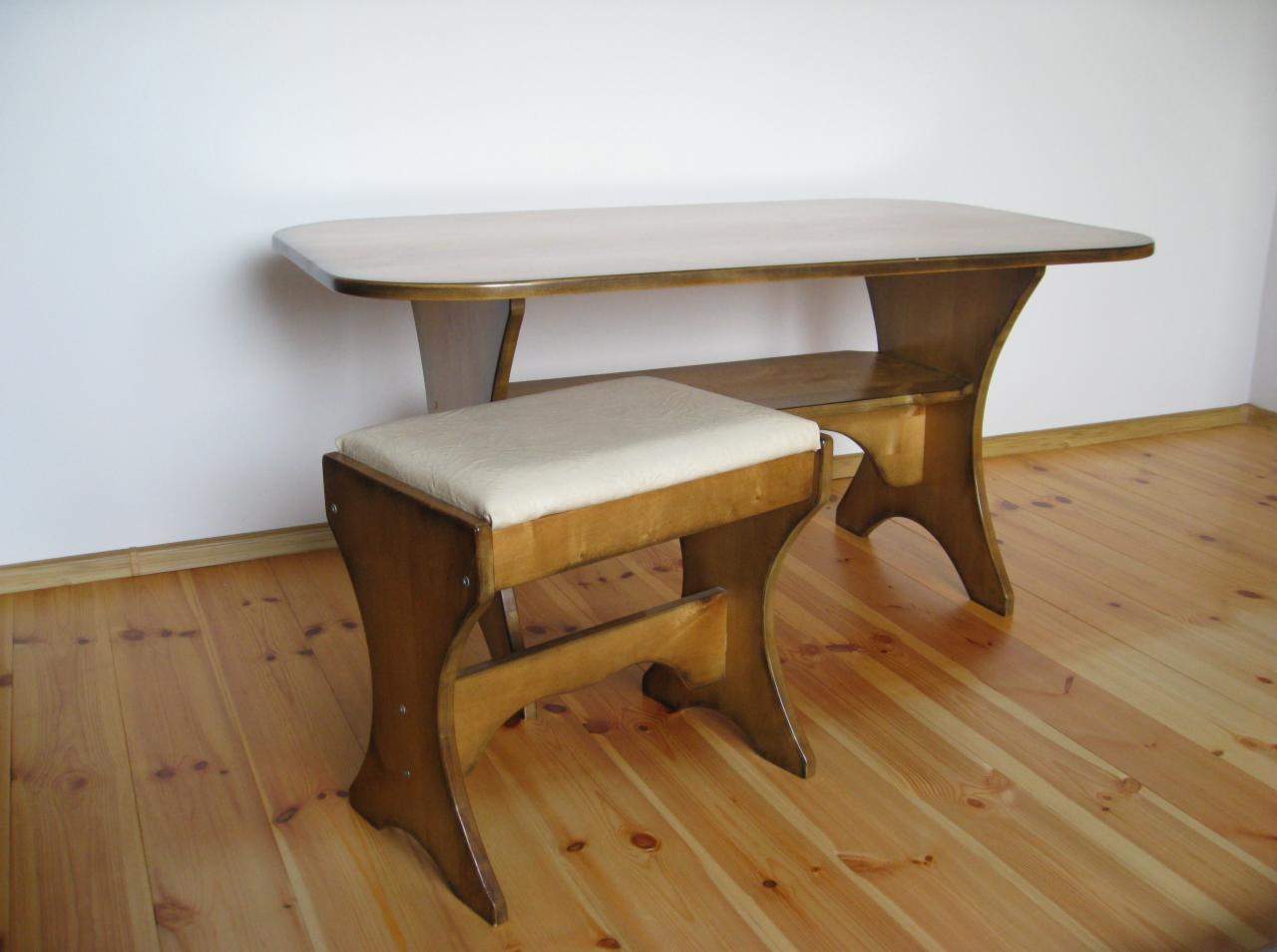 Drewniany stolik z zaokrąglonymi krawędziami i półką pod blatem, w komplecie z tapicerowanym stołkiem na giętych nogach, ustawione na parkiecie w pomieszczeniu z białymi ścianami.