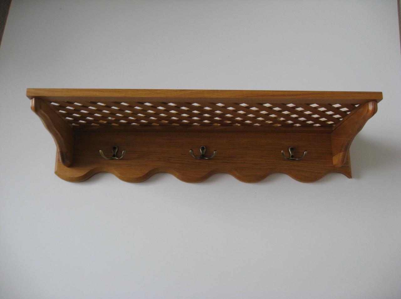 Półka ścienna z drewna dębowego z trzema metalowymi haczykami i ażurową górną częścią, zamontowana na białej ścianie.