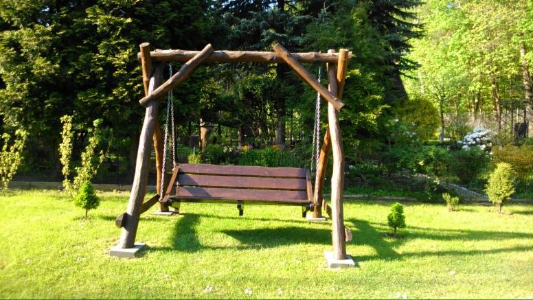 Drewniana huśtawka ogrodowa z siedziskiem z desek, zawieszona na łańcuchach, umieszczona na betonowych podstawach, otoczona zielonym trawnikiem i drzewami.