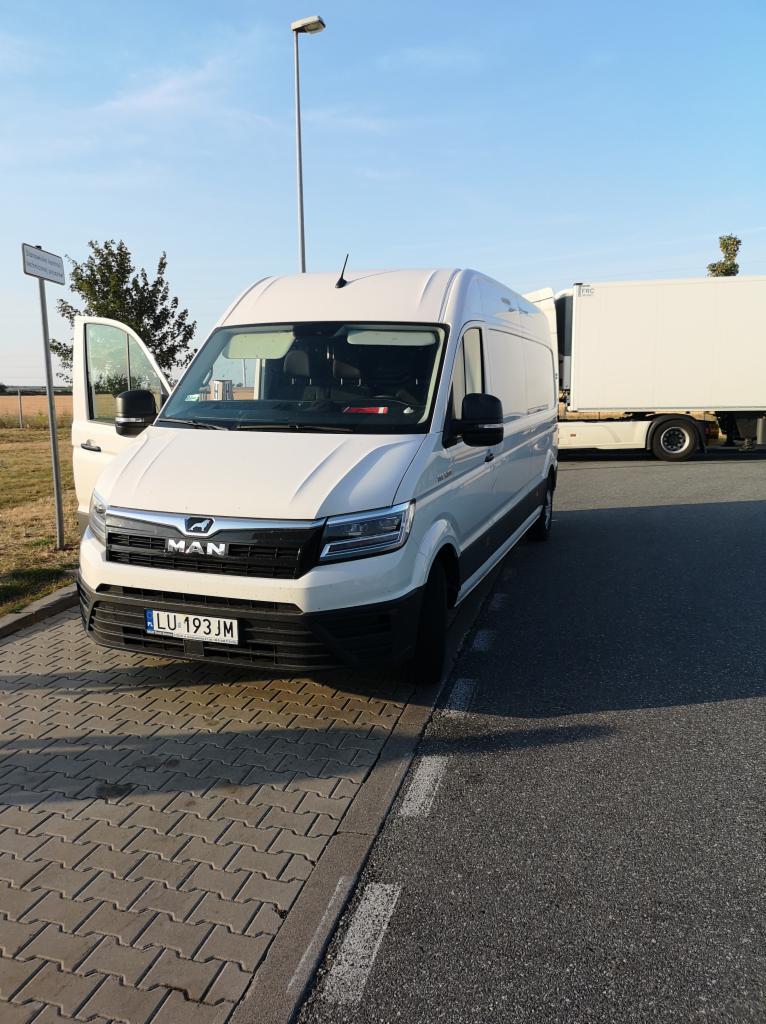 Biały bus MAN z otwartymi drzwiami kierowcy, zaparkowany obok naczepy. Tablica rejestracyjna LU 193JM. Widoczne drzewo i słup oświetleniowy.