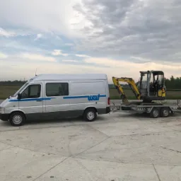 Srebrny bus z niebieskimi pasami ciągnie żółtą mini koparkę na przyczepie, zaparkowany na betonowym placu, w tle pole i pochmurne niebo.