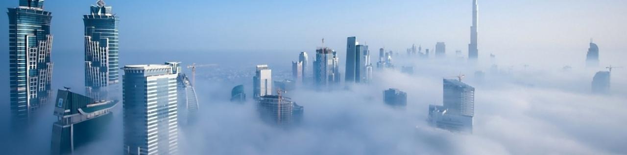 Panorama Dubaju spowita poranną mgłą, widoczne drapacze chmur w trakcie budowy, żurawie budowlane wystające ponad chmury.
