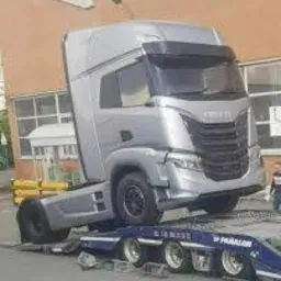 Srebrny ciągnik siodłowy marki Iveco wjeżdża na niskopodwoziową lawetę. Widoczny fragment budynku w tle.