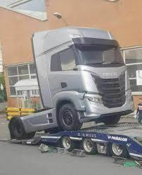 Srebrny ciągnik siodłowy marki Iveco wjeżdża na niskopodwoziową lawetę. Widoczny fragment budynku w tle.