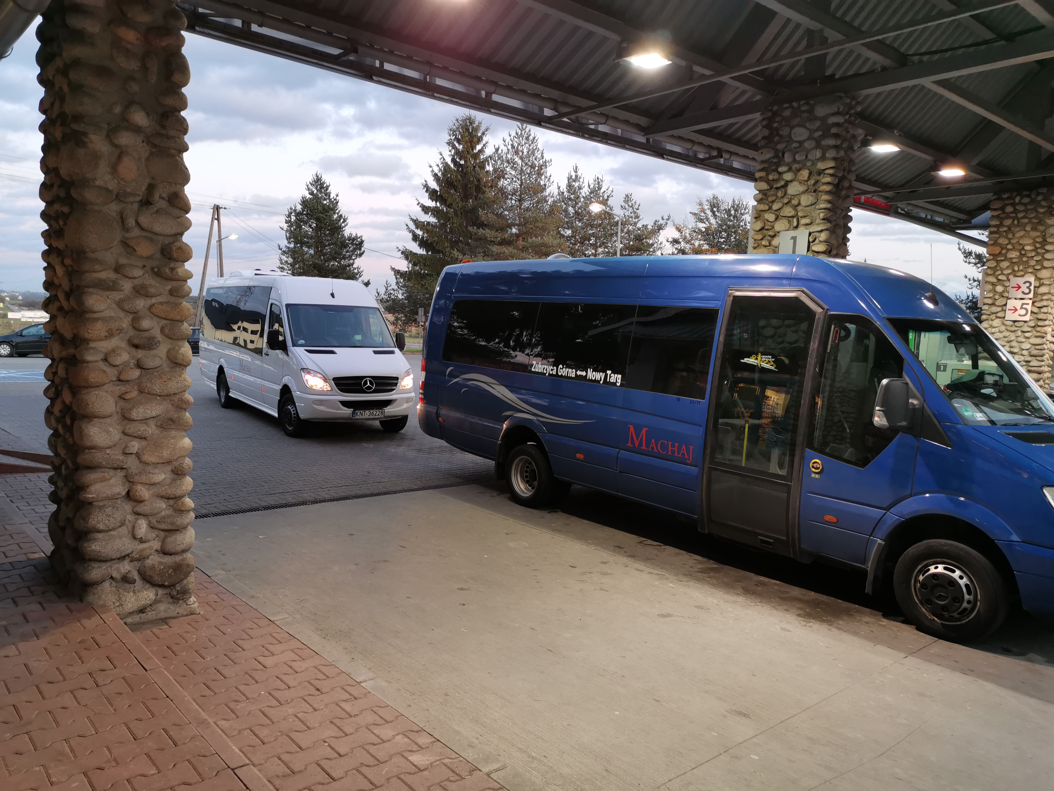 Dwa busy, biały i niebieski, stoją pod wiatą przystankową z kamiennymi kolumnami, z napisem 'Zubrzyca Górna - Nowy Targ' na niebieskim busie.