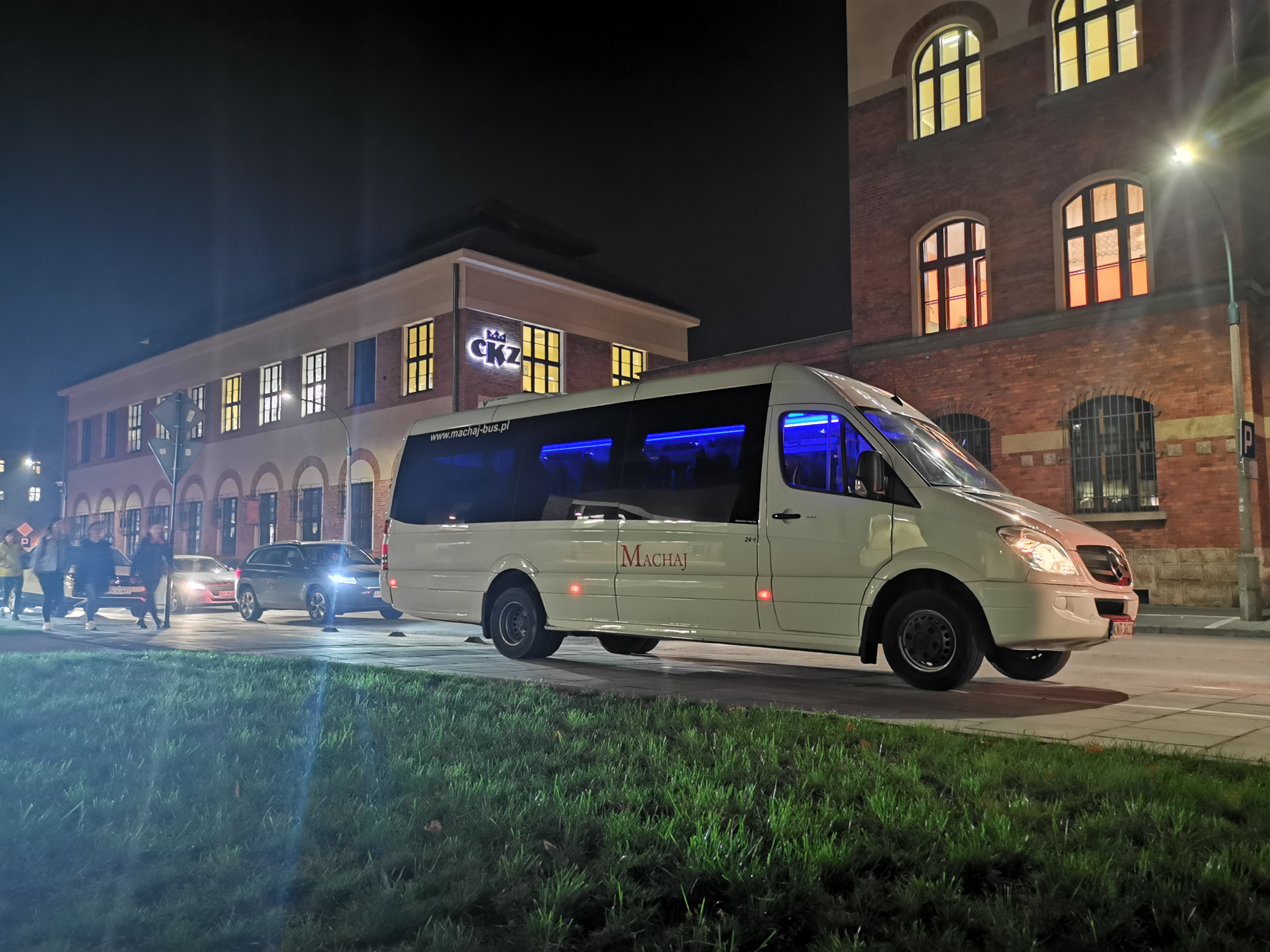 Biały bus marki Mercedes-Benz z niebieskim oświetleniem wnętrza, z napisem 'MACHAJ' i adresem strony internetowej na tle budynku CKZ oświetlonego nocą.