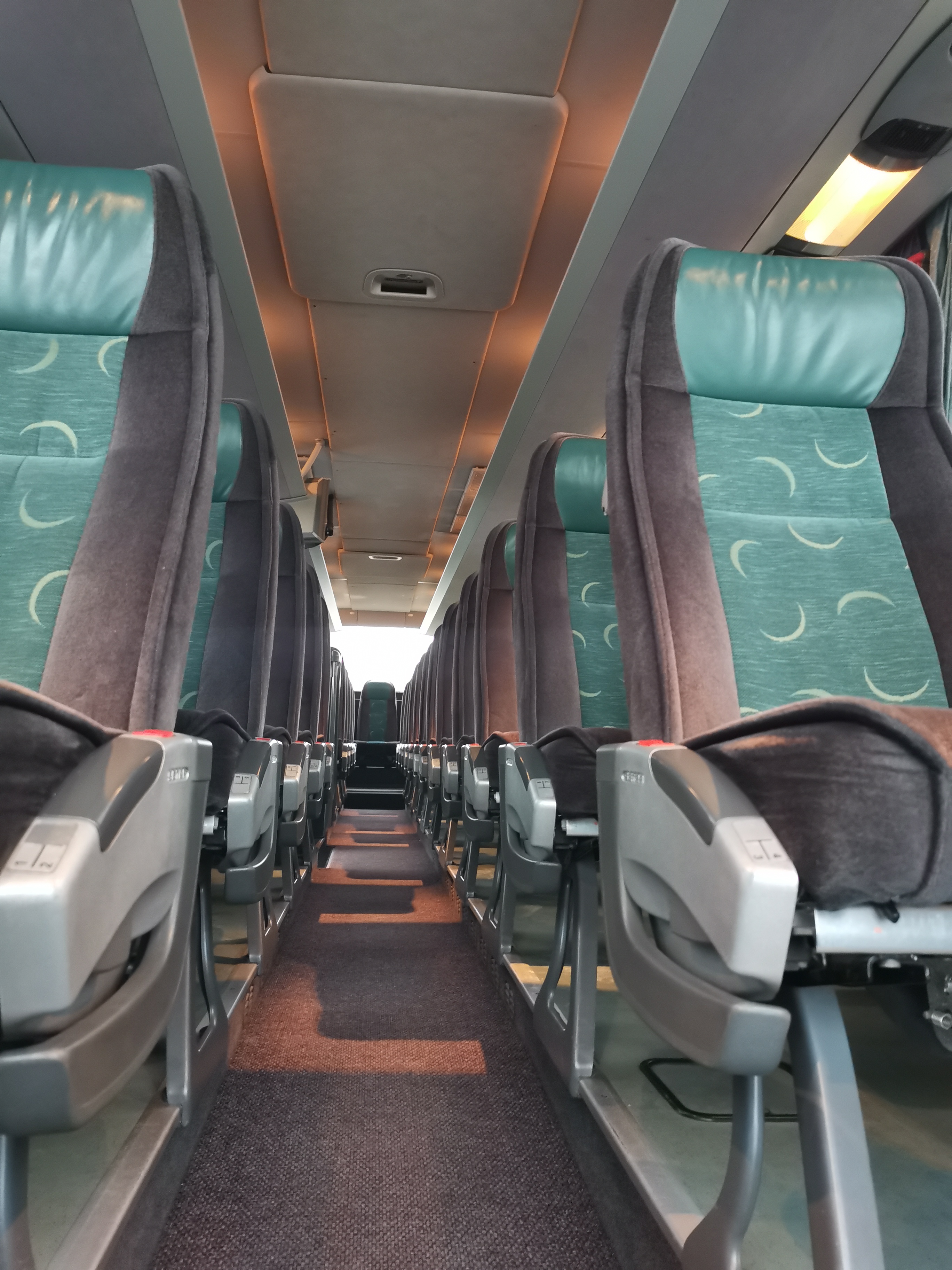 Wnętrze autobusu turystycznego z rzędami foteli w odcieniach szarości i turkusu, widok wzdłuż korytarza z oświetleniem sufitowym.