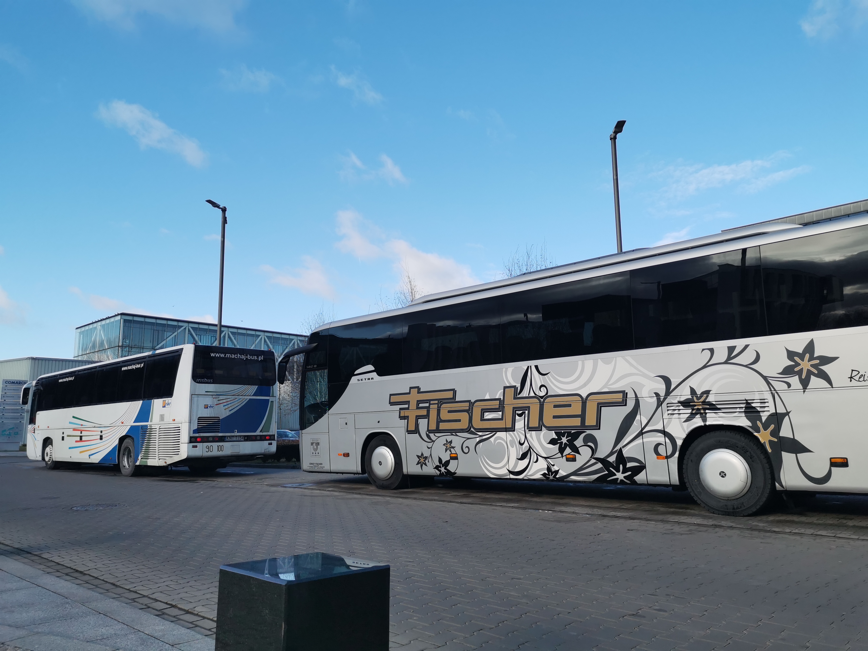 Dwa autokary na parkingu; jeden biało-niebieski z napisem www.machaj-bus.pl, drugi srebrno-biały z dekoracyjnymi wzorami kwiatowymi i napisem Fischer w złotym kolorze.
