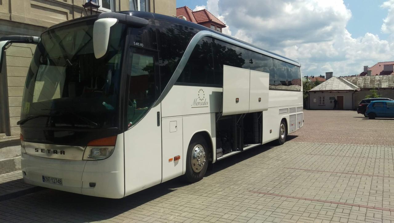 Biały autobus Setra Mercedes z otwartymi lukami bagażowymi zaparkowany na placu z kostki brukowej, widoczne budynki w tle.