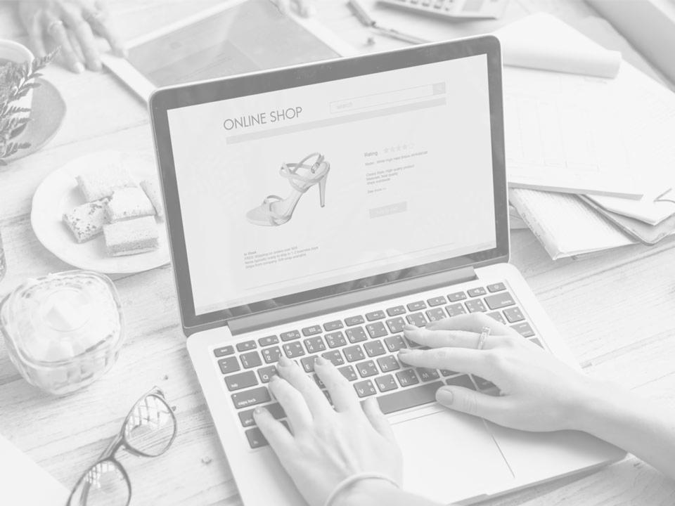 Kobieta wpisuje tekst na laptopie wyświetlającym stronę sklepu internetowego z butami, obok leżą okulary, ciastka i tablet.