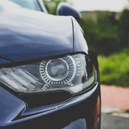 Detaliczny widok reflektora nowoczesnego, ciemnoniebieskiego samochodu na tle zieleni i czerwonej kostki brukowej.