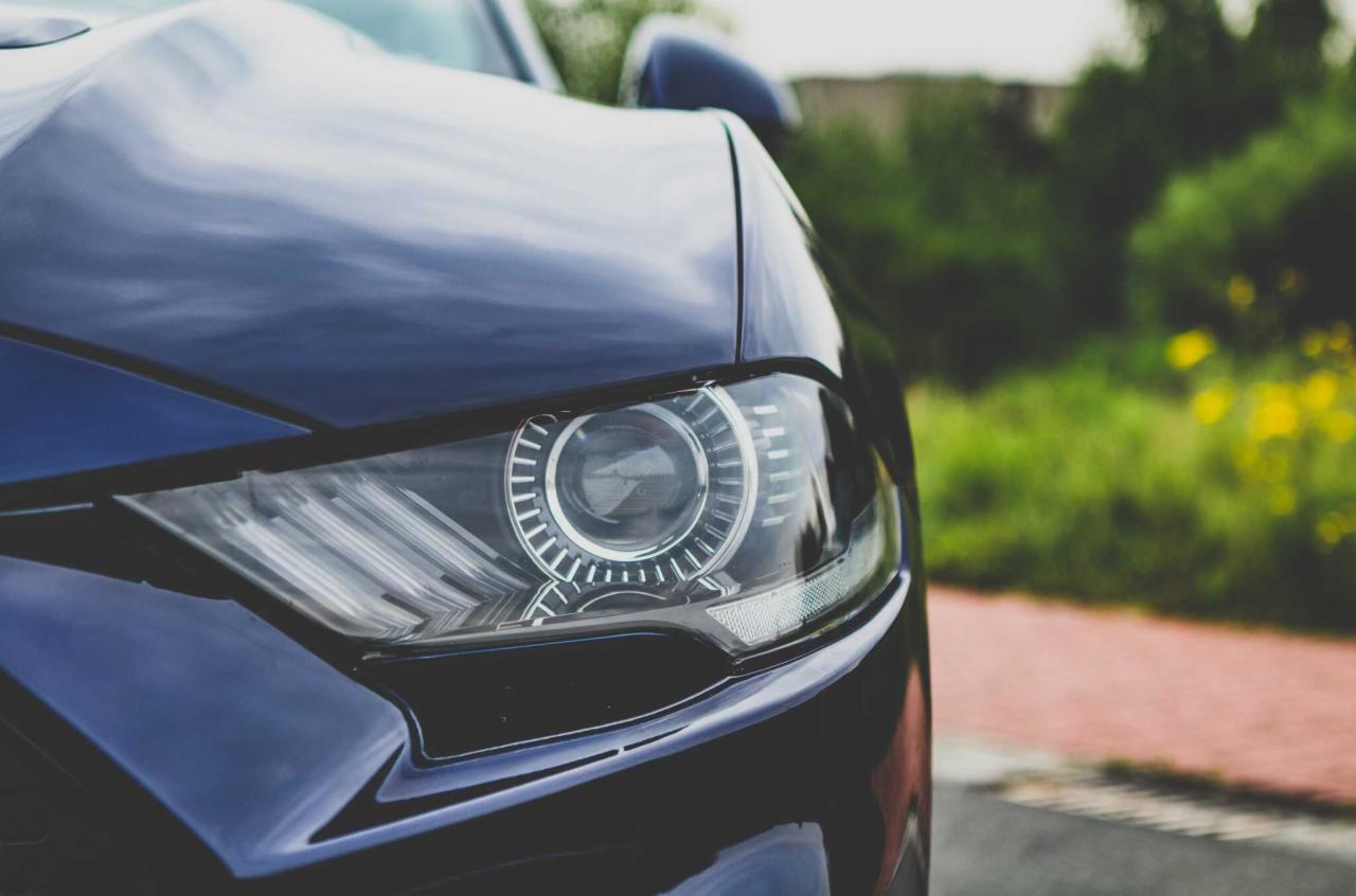 Detaliczny widok reflektora nowoczesnego, ciemnoniebieskiego samochodu na tle zieleni i czerwonej kostki brukowej.
