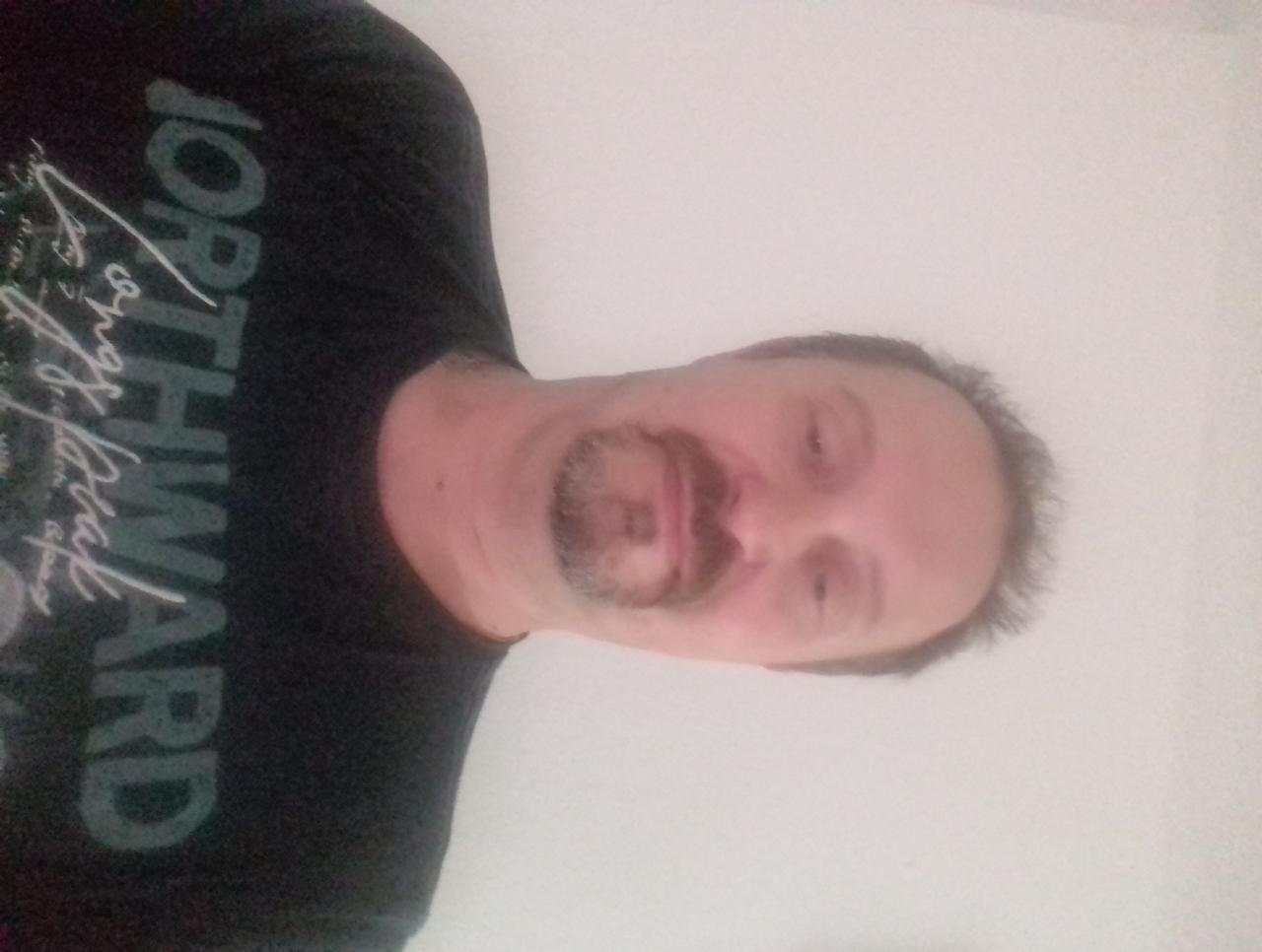 Portret mężczyzny z krótką brodą i wąsami, ubranego w ciemną koszulkę z białym napisem 'NORTHWARD'. Białe tło. Zdjęcie obrócone.