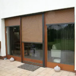 Brązowe rolety zewnętrzne częściowo opuszczone na dużych, przesuwnych oknach tarasowych z drewnianymi ramami, widok z zewnątrz na taras wyłożony płytkami.