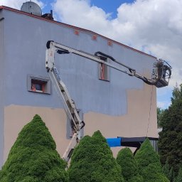 STAEM Michał Kaźmierski - Podnośnik koszowy OIL&STEEL przy elewacji budynku, gdzie trwa malowanie na jasnoszary kolor, widoczny fragment beżowej ściany. Na dachu antena satelitarna, w tle zielone tuje.
