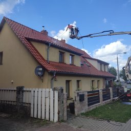 STAEM Michał Kaźmierski - Pracownik na podnośniku koszowym typu pająk myje czerwoną dachówkę domu jednorodzinnego z beżową elewacją, używając myjki ciśnieniowej. Widoczne niebo z chmurami.