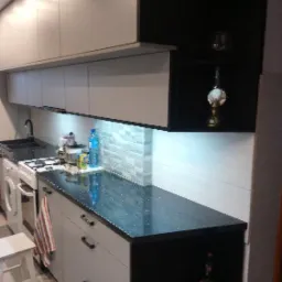 Nowoczesna kuchnia na wymiar z szarymi szafkami górnymi i dolnymi, czarnym blatem z połyskiem i otwartymi czarnymi półkami bocznymi, uzupełniona oświetleniem podszafkowym i dekoracyjną mozaiką...