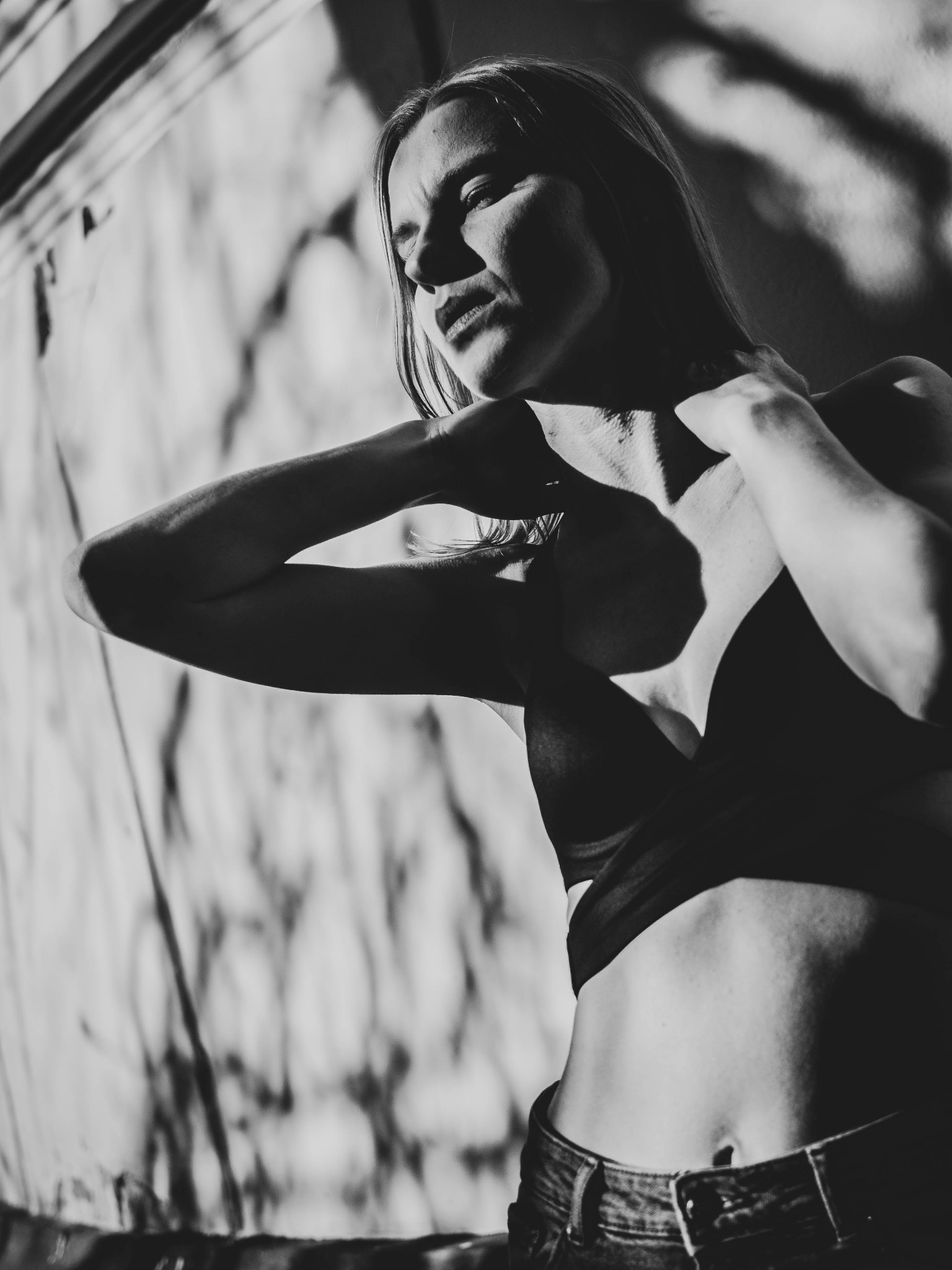 Czarno-białe zdjęcie kobiety w bieliźnie i dżinsach, pozującej na tle ściany z ciekawym cieniem, oświetlonej ostrym światłem.