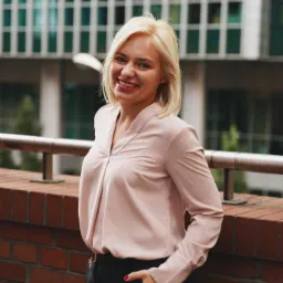 Uśmiechnięta blondynka w różowej bluzce i czarnych spodniach, oparta o ceglany murek na tle nowoczesnego biurowca.