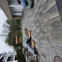 Ułożona, szara kostka brukowa w dwóch wzorach przed nowoczesnym budynkiem, z paletami i narzędziami przygotowanymi do dalszych prac.