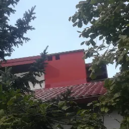 Czerwony budynek z dachem z czerwonej dachówki, widoczny zza drzew, częściowo przysłonięty liśćmi i gałęziami.