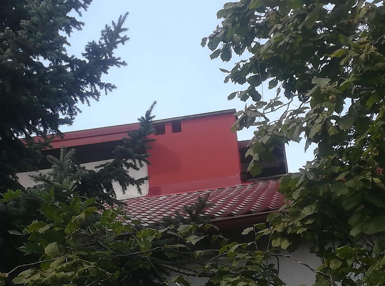 Czerwony budynek z dachem z czerwonej dachówki, widoczny zza drzew, częściowo przysłonięty liśćmi i gałęziami.