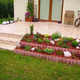 Ogród z tarasem: rabata kwiatowa z obrzeżem z palików, trawnik, schody z kostki, meble ogrodowe przy domu. Aranżacja z lampkami solarnymi.