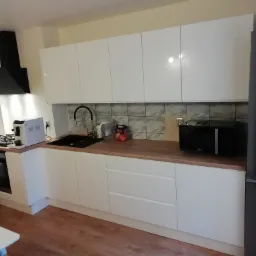 Nowoczesna, jasna kuchnia z białymi, błyszczącymi frontami szafek, drewnianym blatem, czarnym okapem i czarnym zlewem. Na blacie widoczny ekspres do kawy, mikrofalówka i akcesoria kuchenne.